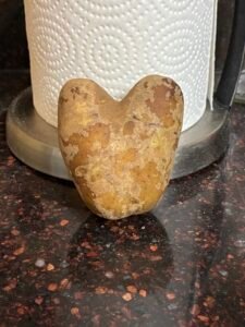Front and center is a real russet potato that has grown in the shape of a heart. It is leaning against a roll of paper towel that is sitting on a counter.